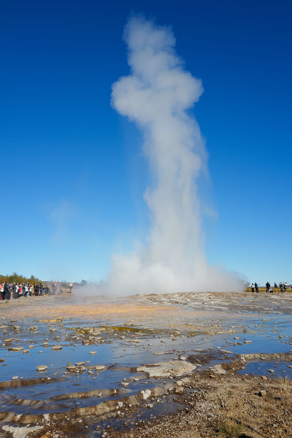 Strokkur geyser erupts with a tall column of steam and water against bright blue sky in Iceland's geothermal area.