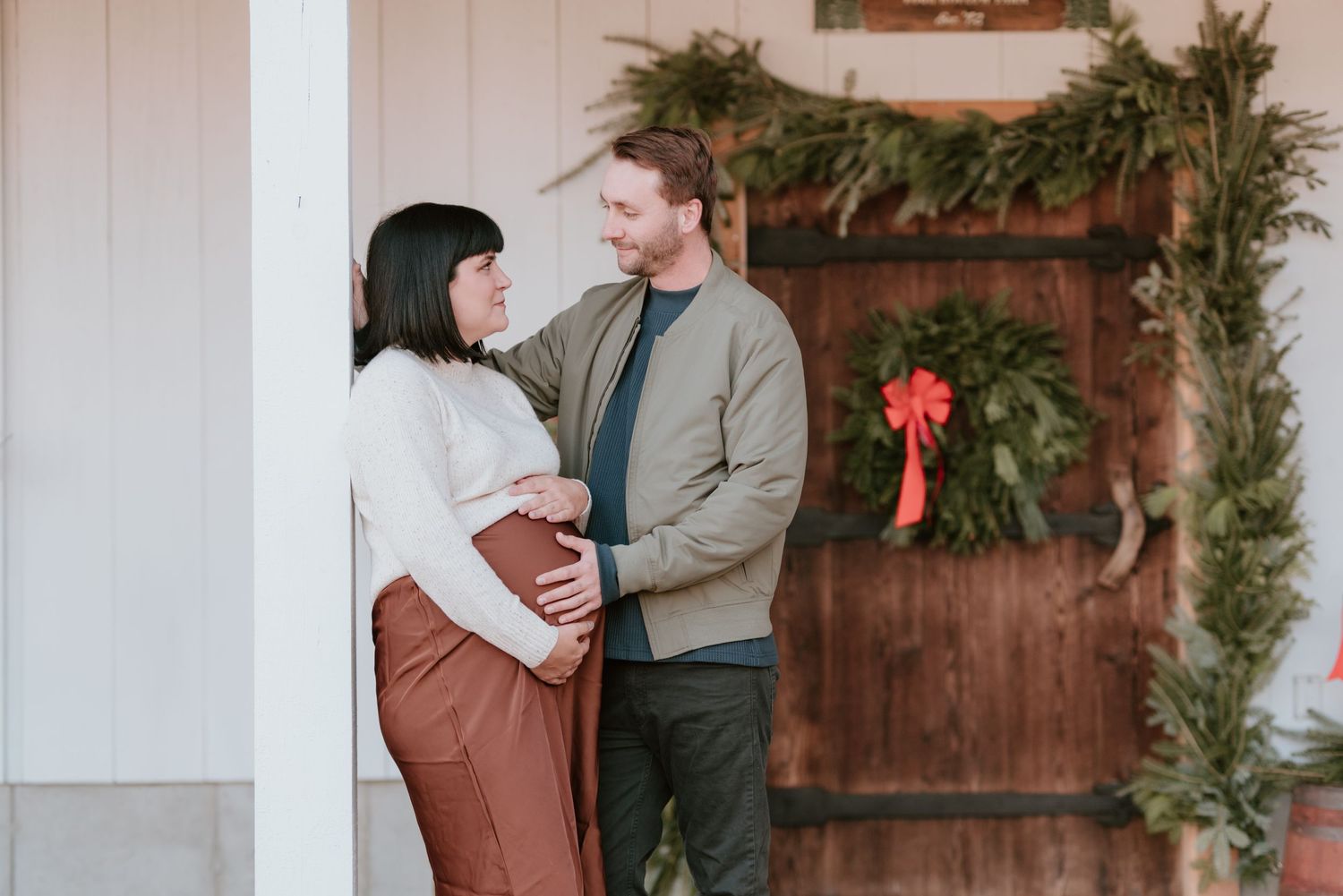 Couple kissing while holding hands among rows of green Christmas trees at a tree farm.