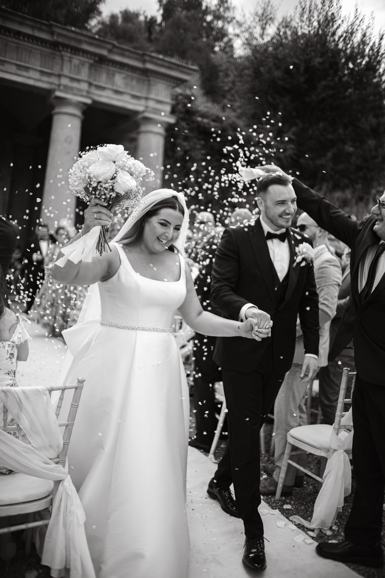 Newlyweds exit their ceremony under a shower of confetti at an outdoor venue with columns.