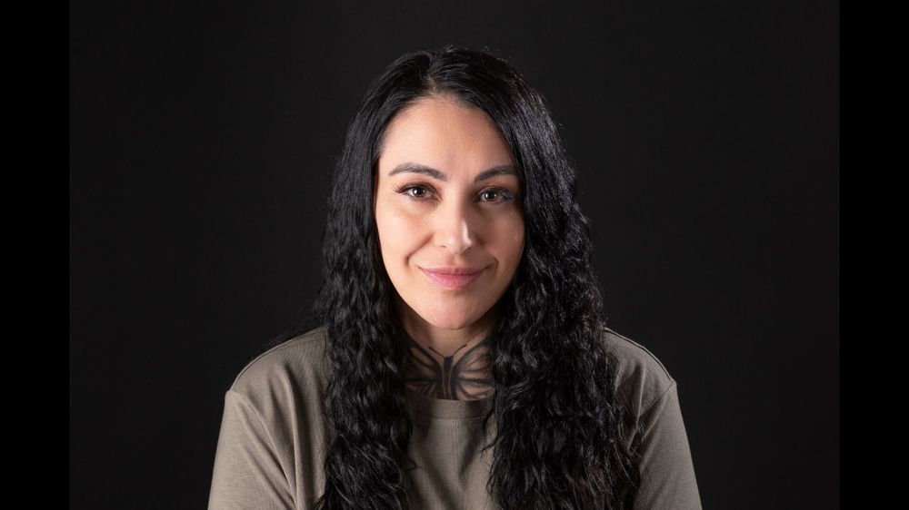 Portrait with long dark curly hair wearing taupe hoodie against black background.