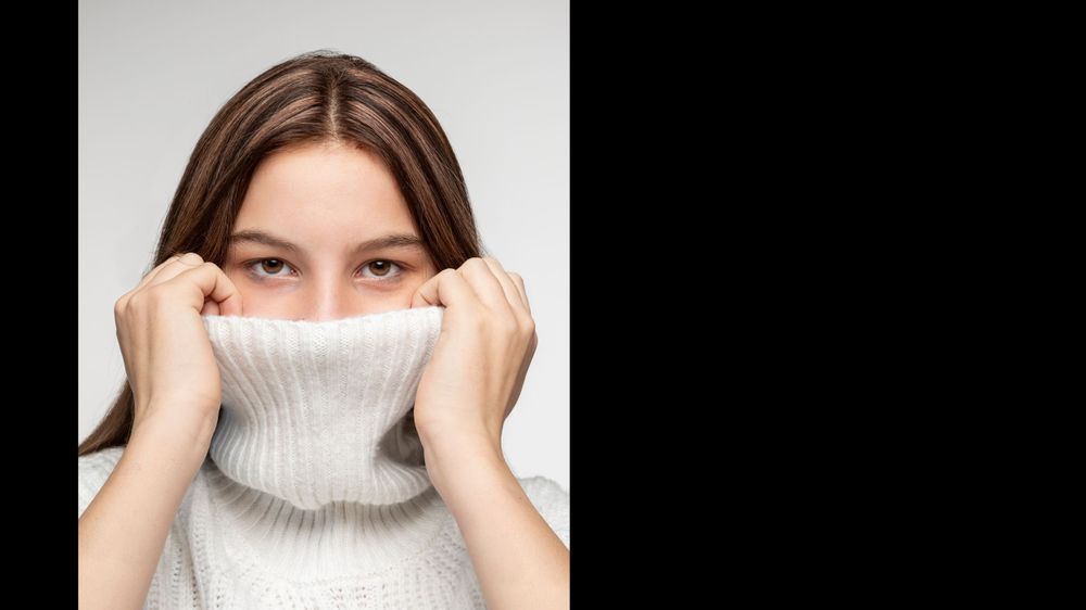 Person pulling white turtleneck sweater up to cover face against white background.