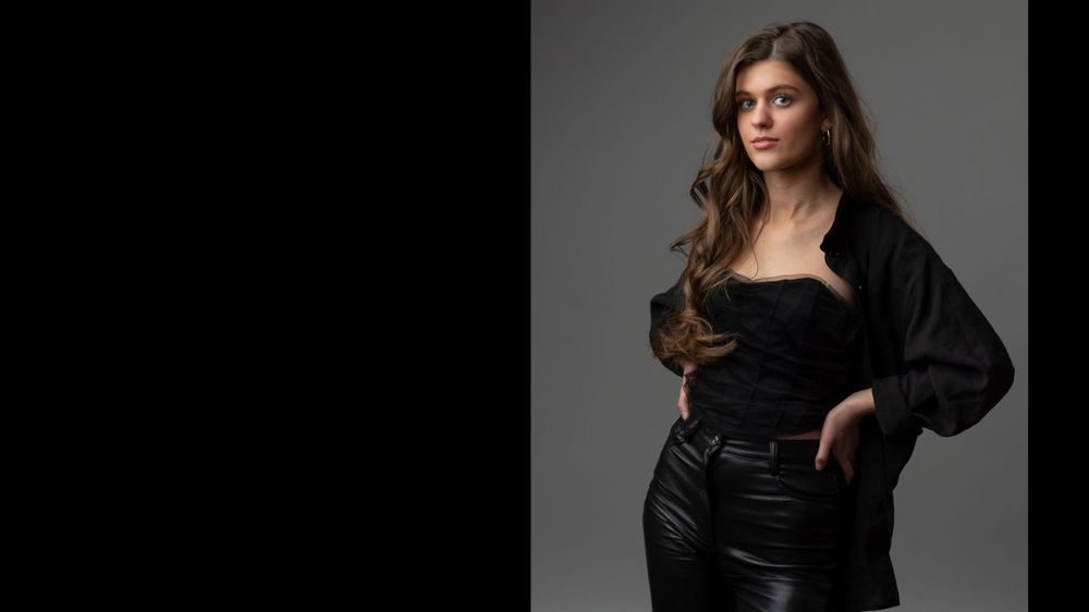 A dramatic fashion portrait in black attire against a dark background with moody lighting.
