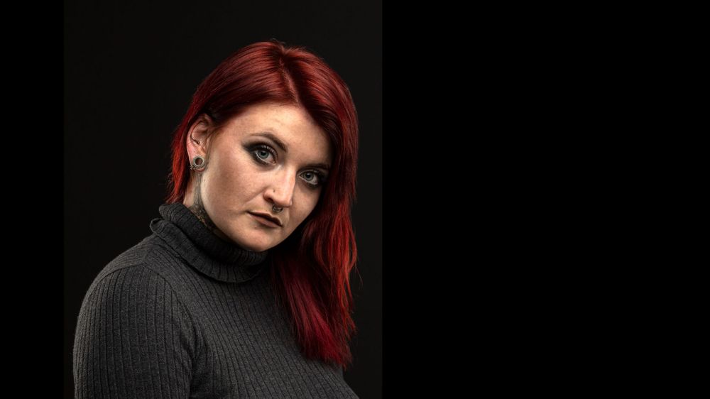 Portrait of a person with vibrant red hair wearing a dark turtleneck against a black background.
