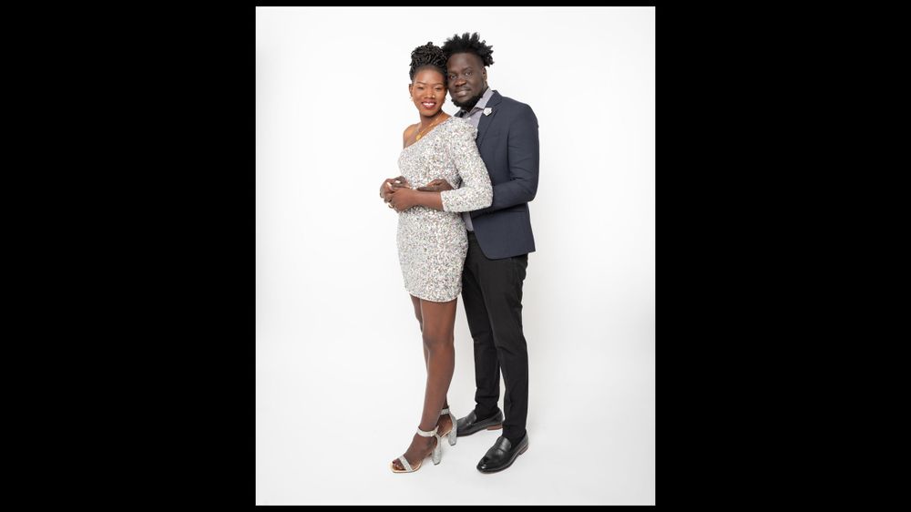 Elegant couple in formal wear, one in white dress and one in black suit, embracing against white background.