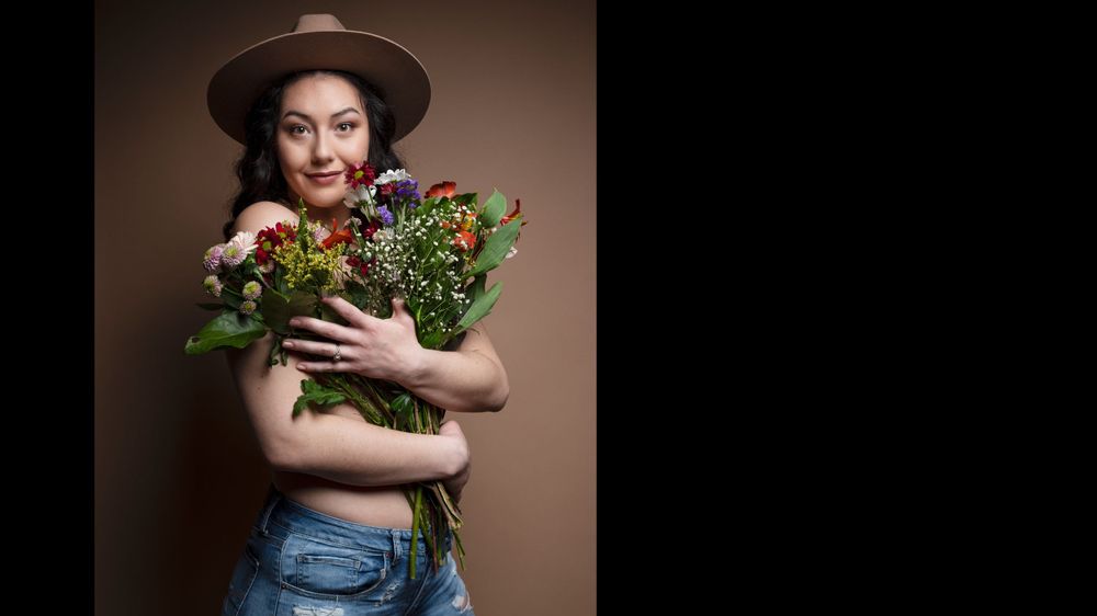 Person in hat and denim holding colorful flowers against brown backdrop.