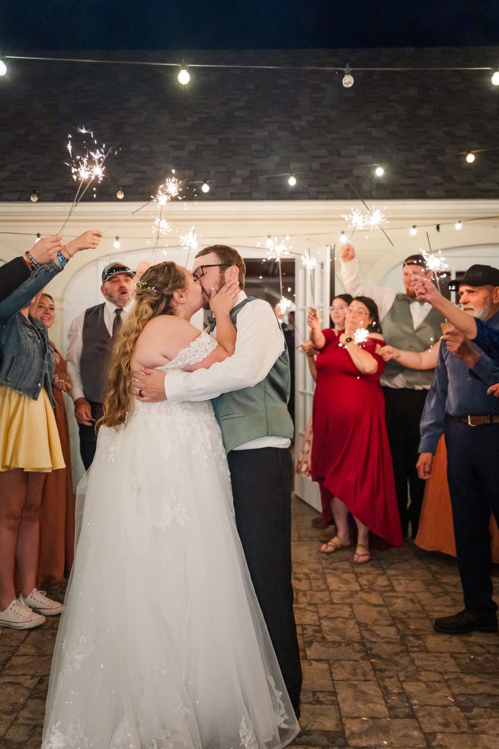 A romantic sparkler-lit kiss during wedding reception with guests celebrating in background.