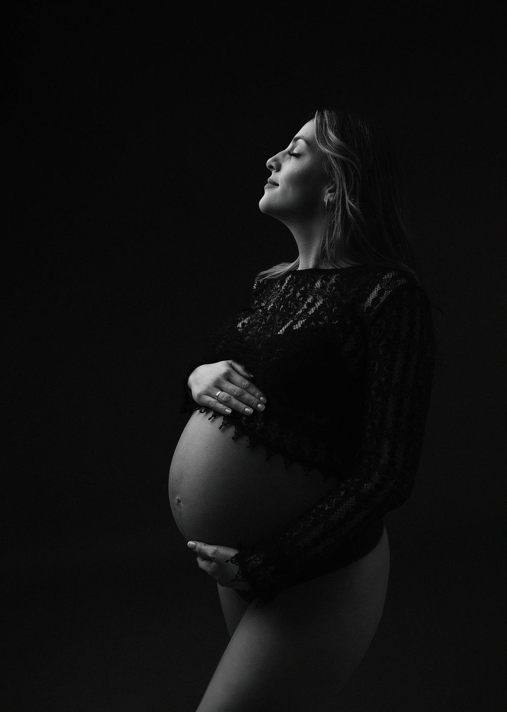 Dramatic black and white portrait of pregnant woman in lace dress against dark background.