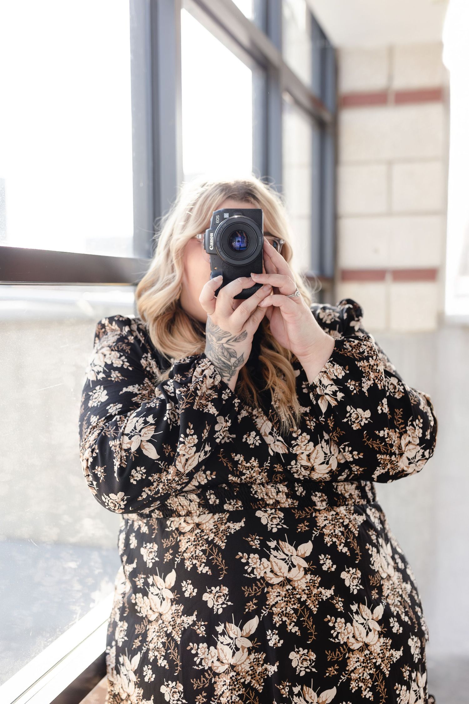 Photographer in floral dress takes photos near large windows in bright indoor space.