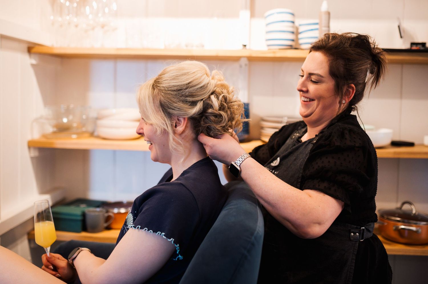 A hairstylist wearing black works on styling a client's blonde hair in a salon setting with wooden shelves in background.
