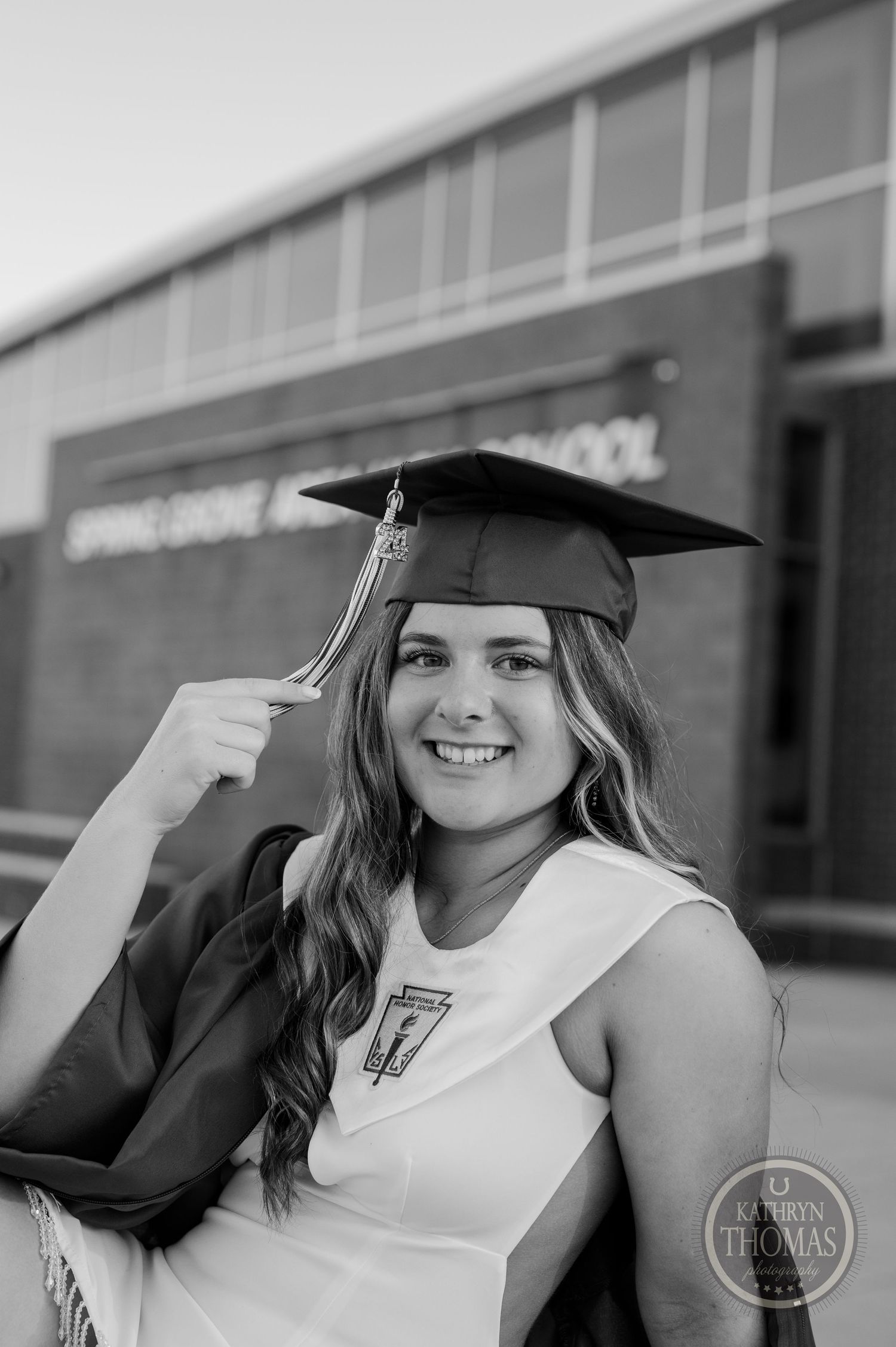 Graduate wearing black cap and gown poses in front of Spring Grove High School.