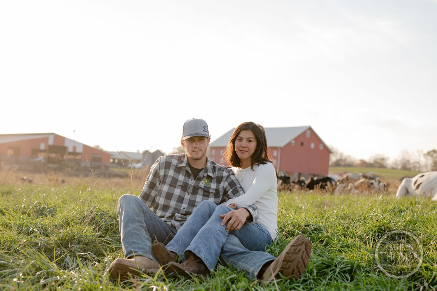 A couple sits together in a grassy field with a red barn and farm animals in the background.