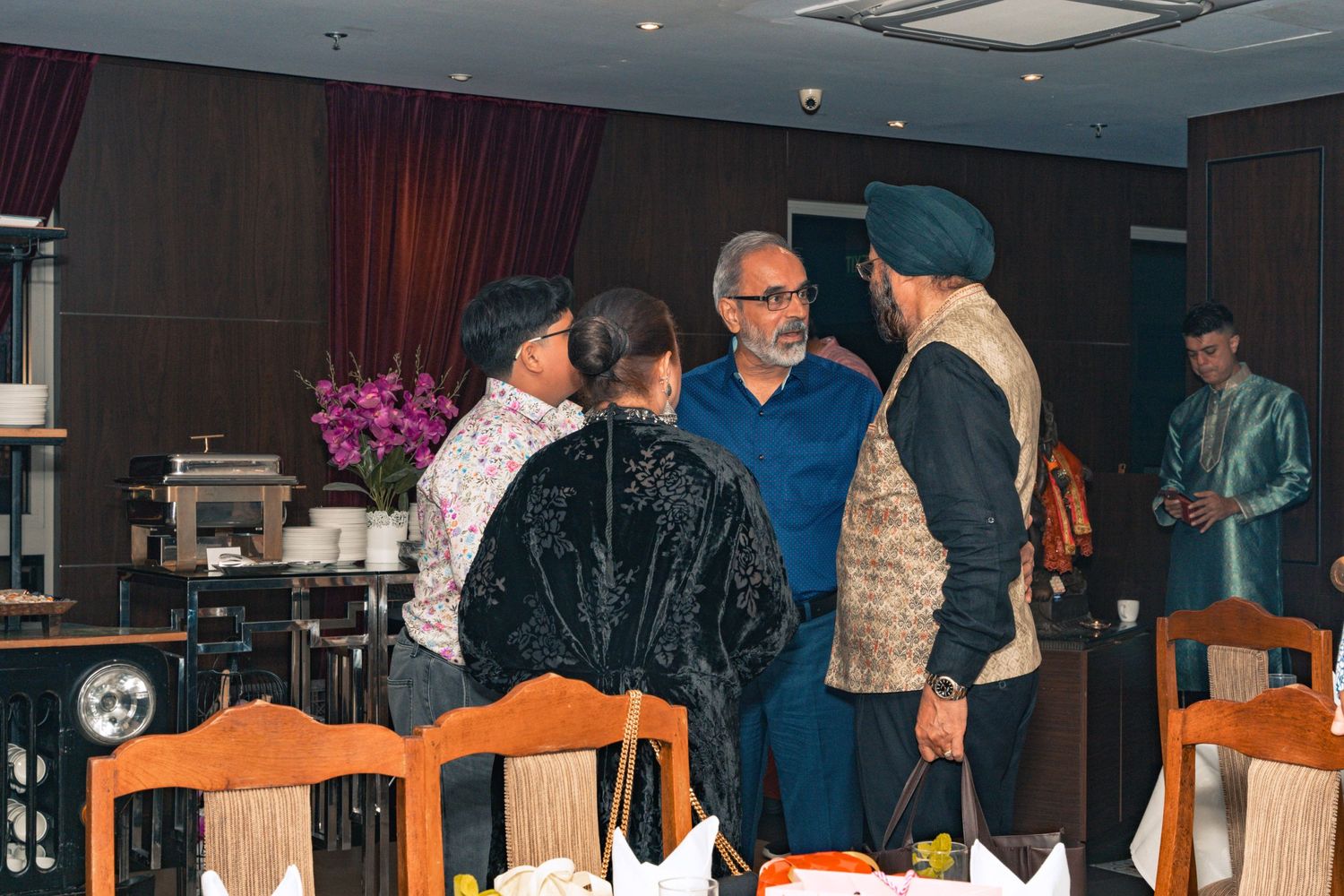 Group engaged in conversation at an indoor dining event with orange chairs and dark curtains.