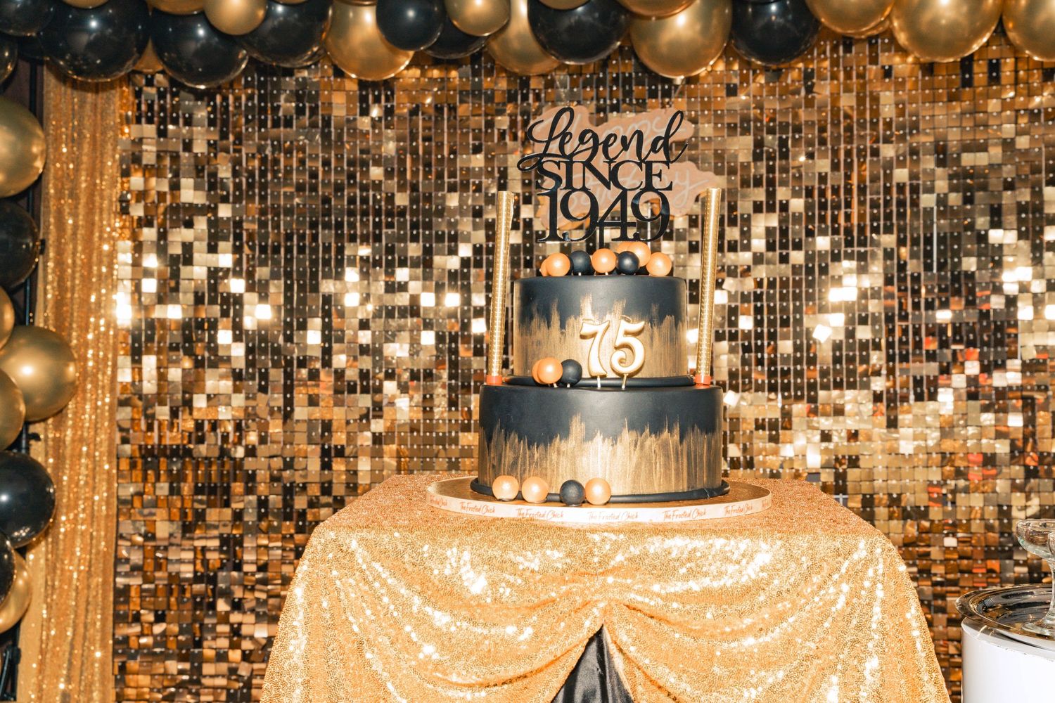 Elegant black and gold celebration cake display with sequined backdrop and balloon decorations.