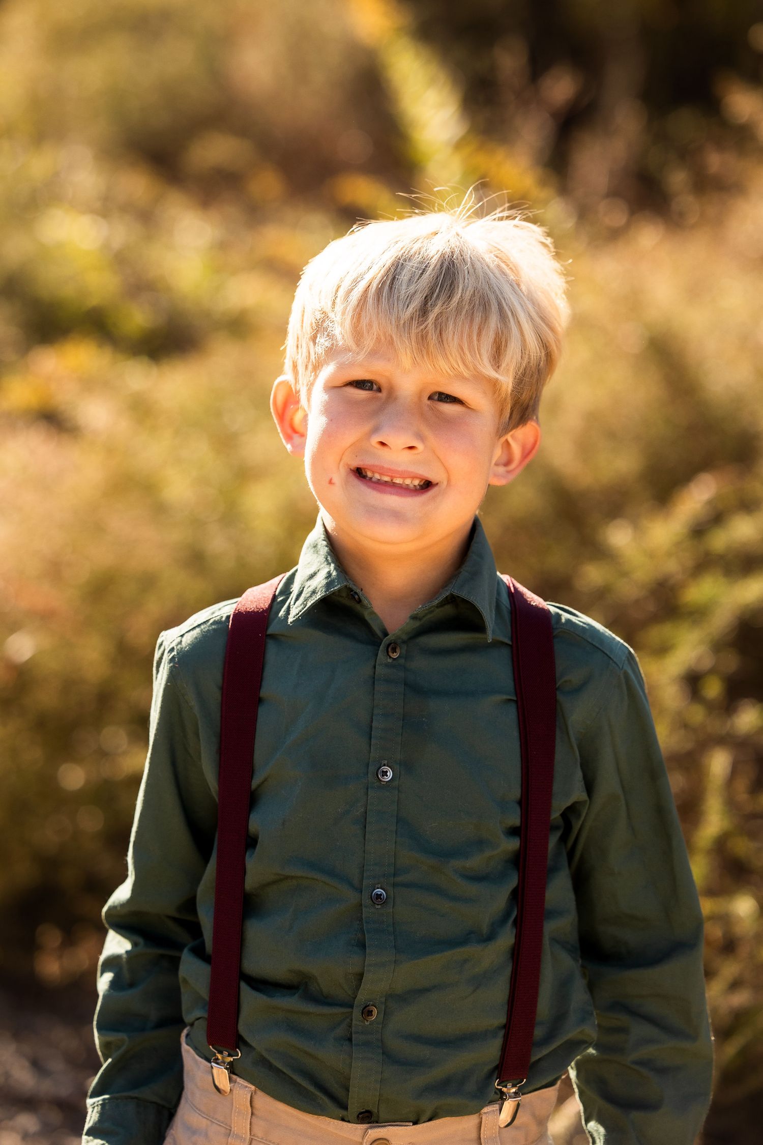 Child wearing green shirt and suspenders smiles during autumn photo session.