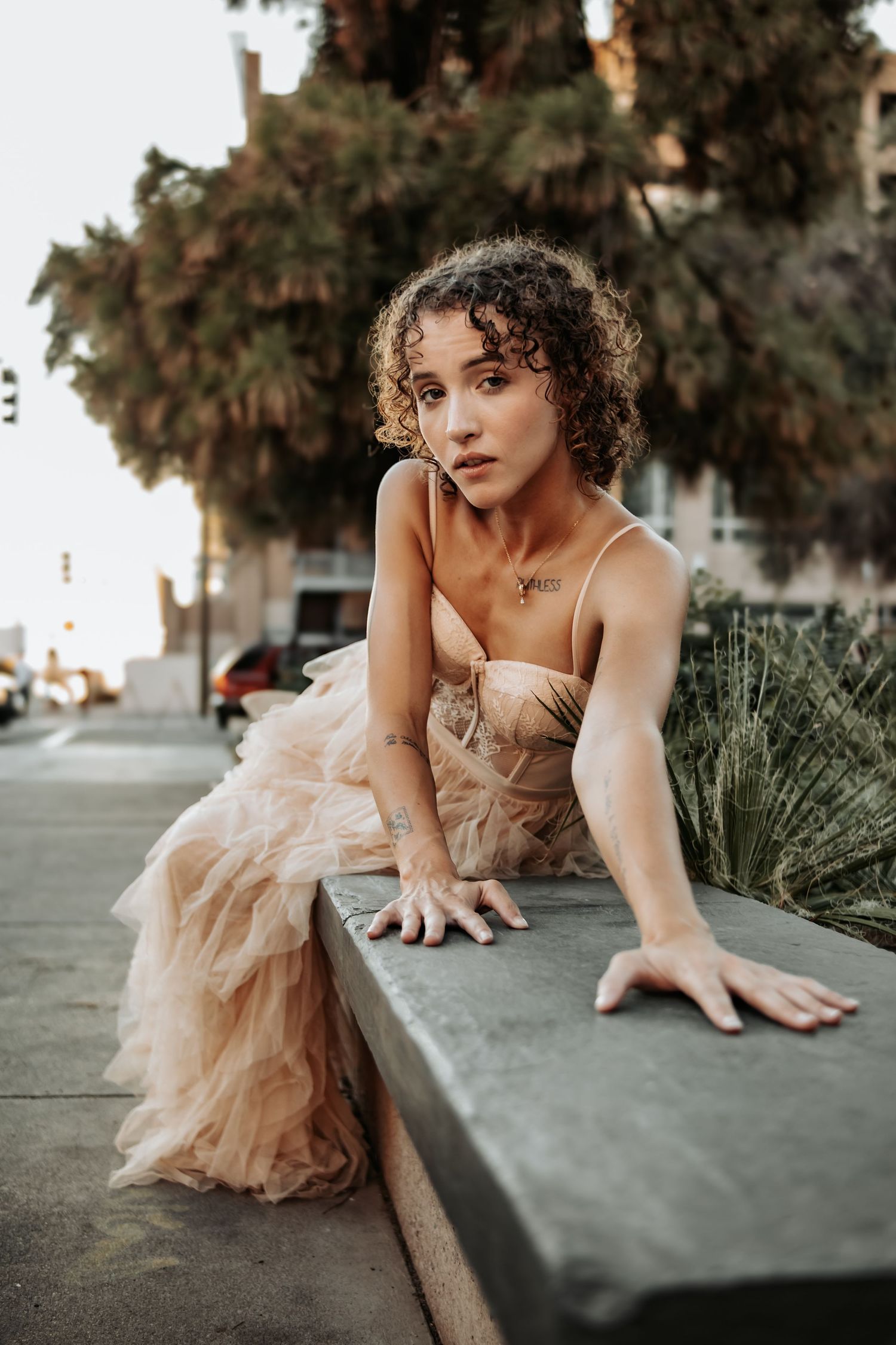 Portrait in romantic peach dress sitting on concrete ledge with urban background.