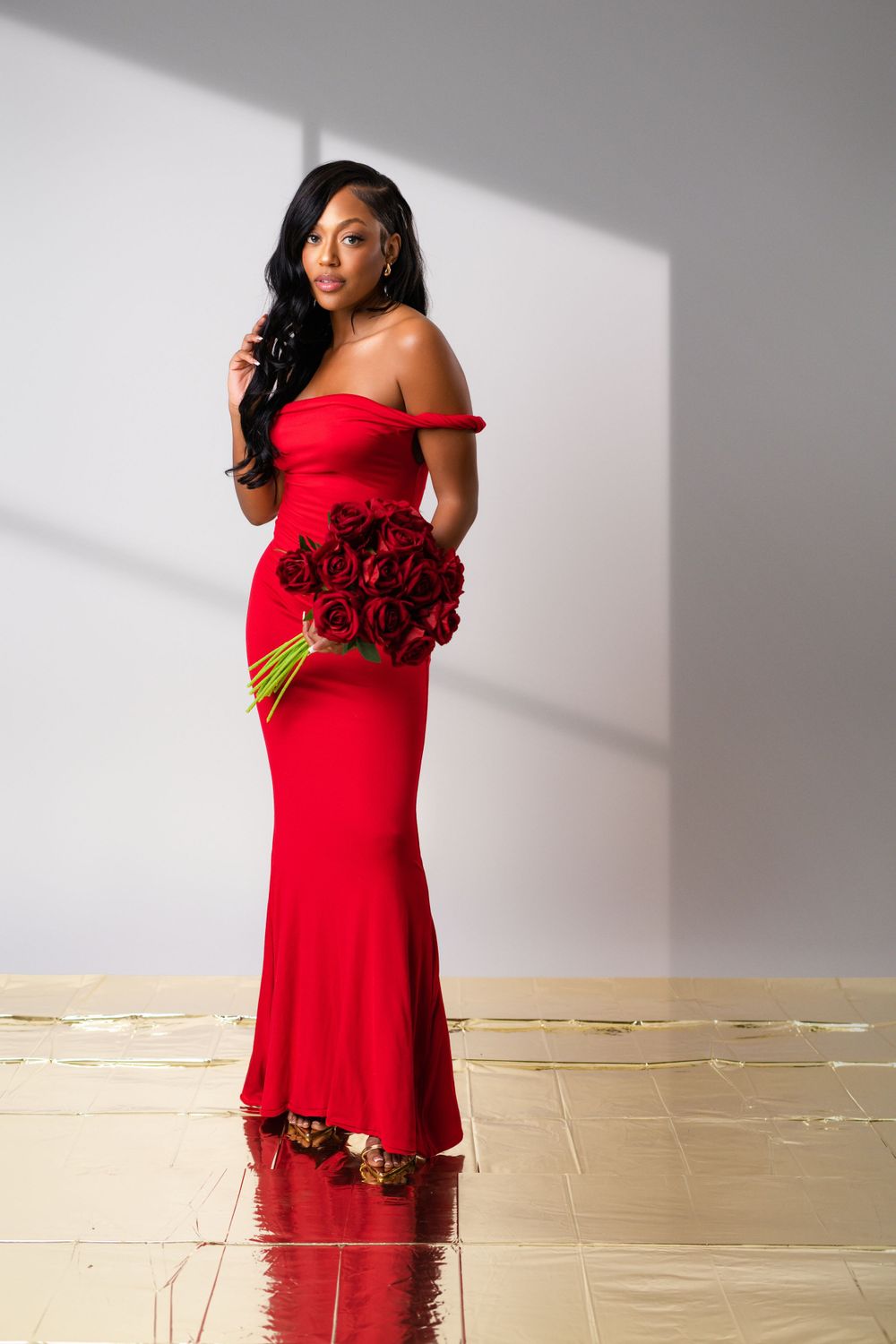 Person in elegant red off-shoulder mermaid gown holds bouquet of red roses against white wall with dramatic lighting.