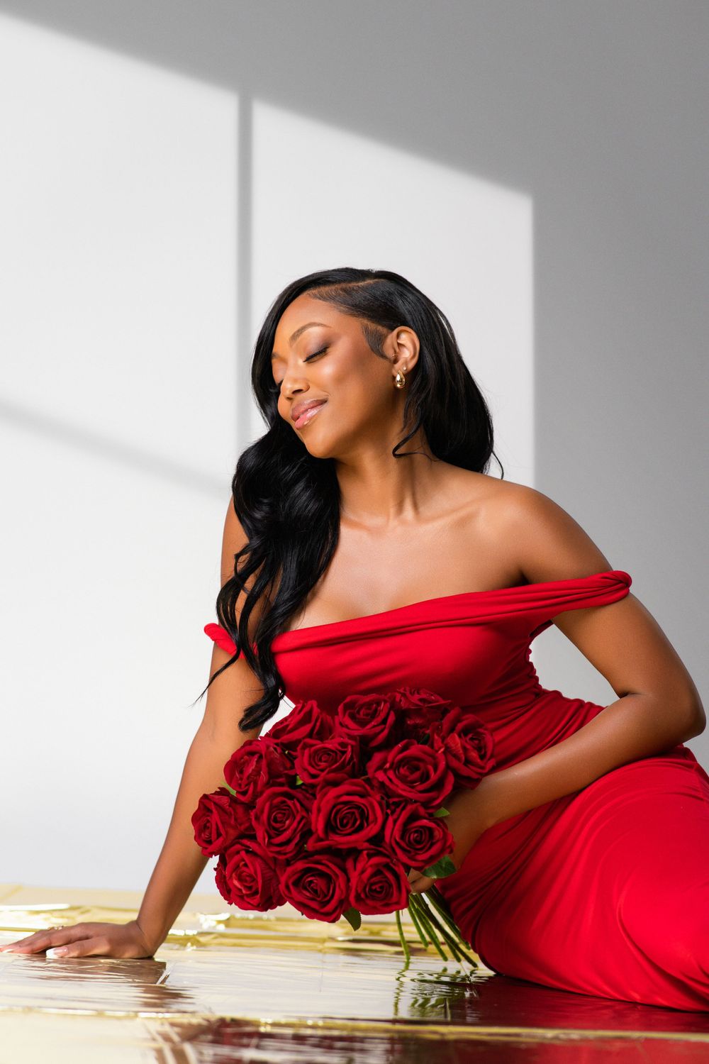 A person in an elegant off-shoulder red dress poses with a bouquet of red roses in dramatic window lighting.