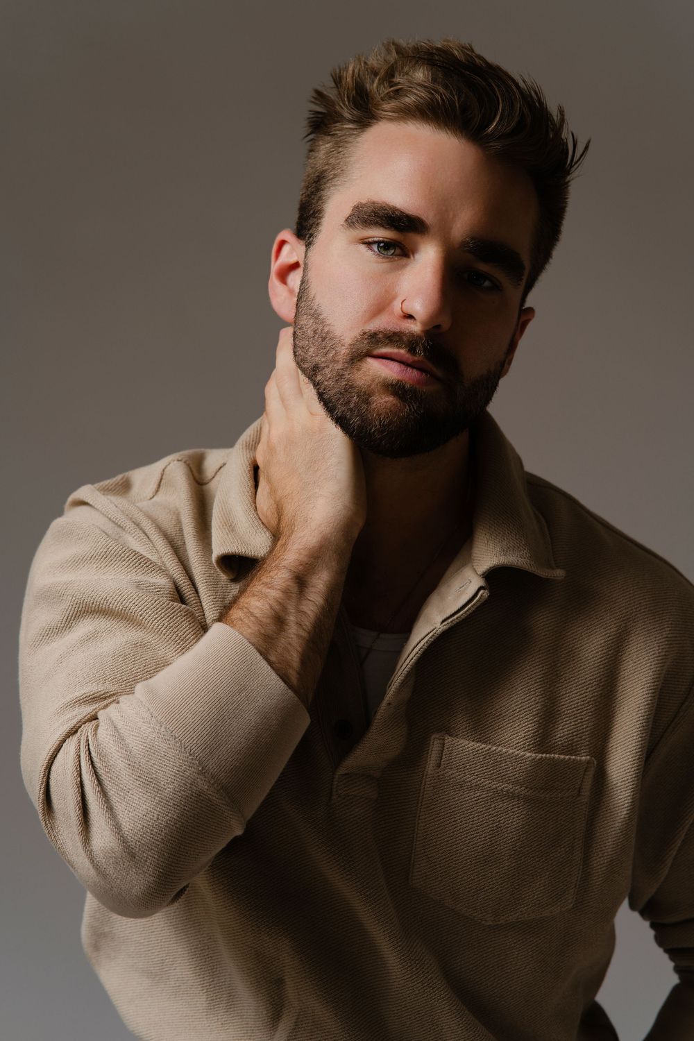 A man in a beige jacket poses against a neutral background with his hand thoughtfully placed on his neck.