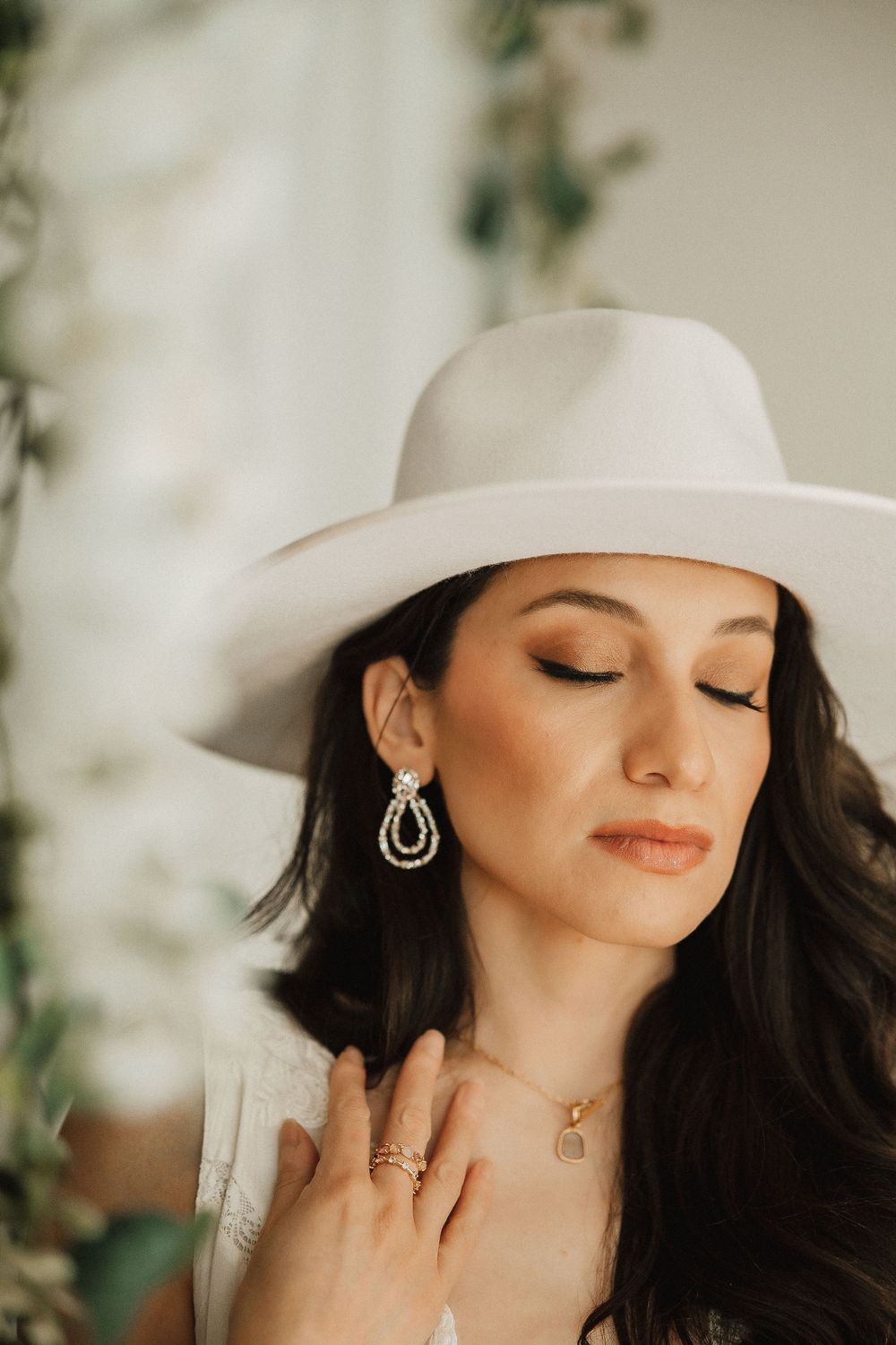 A close up shot of someone in a white cowboy hat with long dark hair wearing dangling earrings and delicate jewelry.