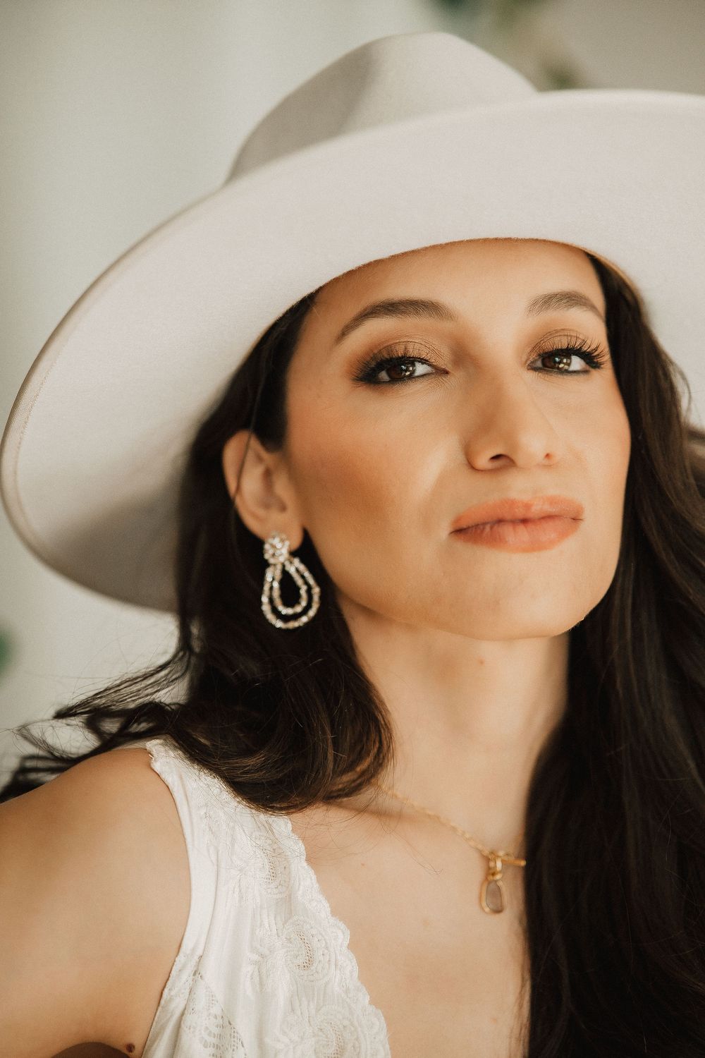 A close-up portrait featuring someone wearing a white cowboy hat, white lace top and sparkling teardrop earrings.