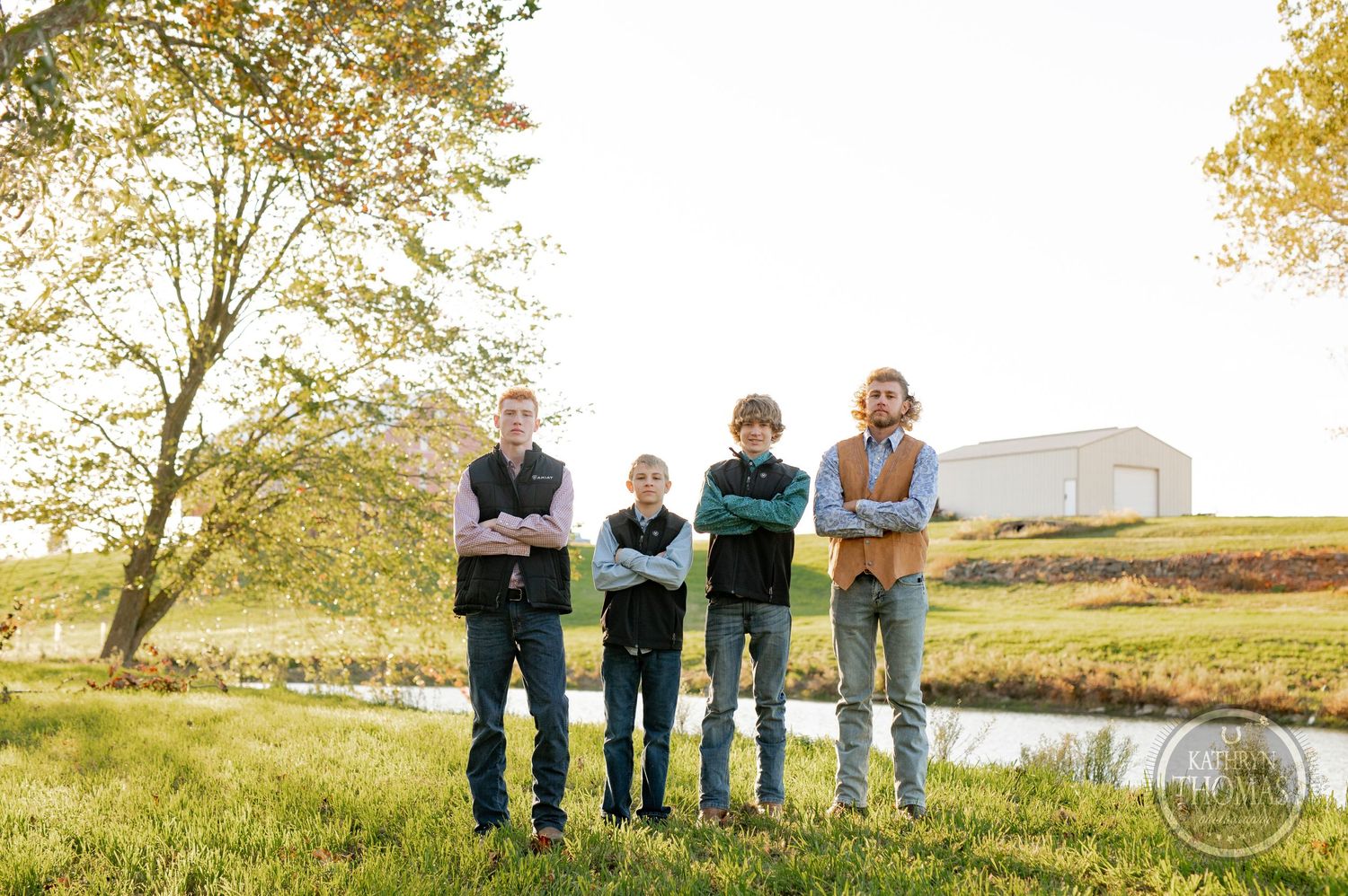 Four people standing in a row on grassy field with pond and building in background.