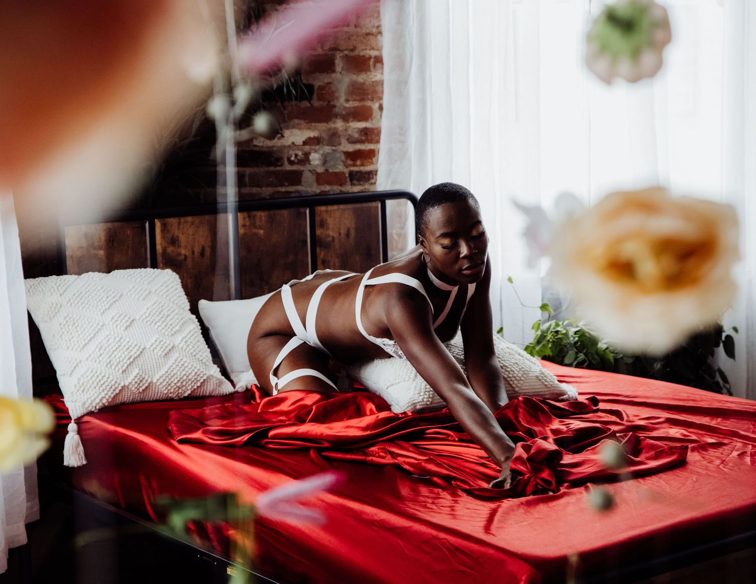 A person poses on a bed with red satin sheets in a bedroom with exposed brick walls and natural lighting from windows.