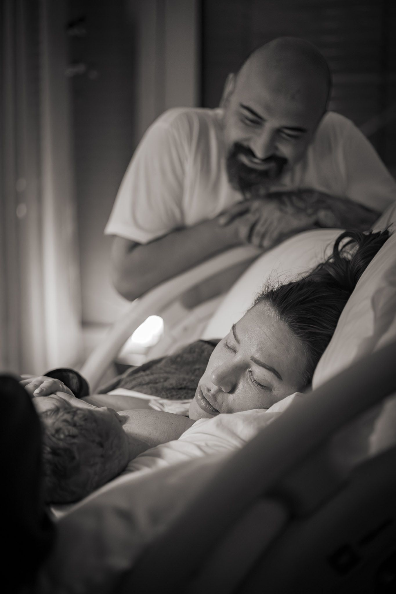 Svartvitt foto på en nyförlöst kvinna med sitt barn på bröstet. Pappan står i bakgrunden