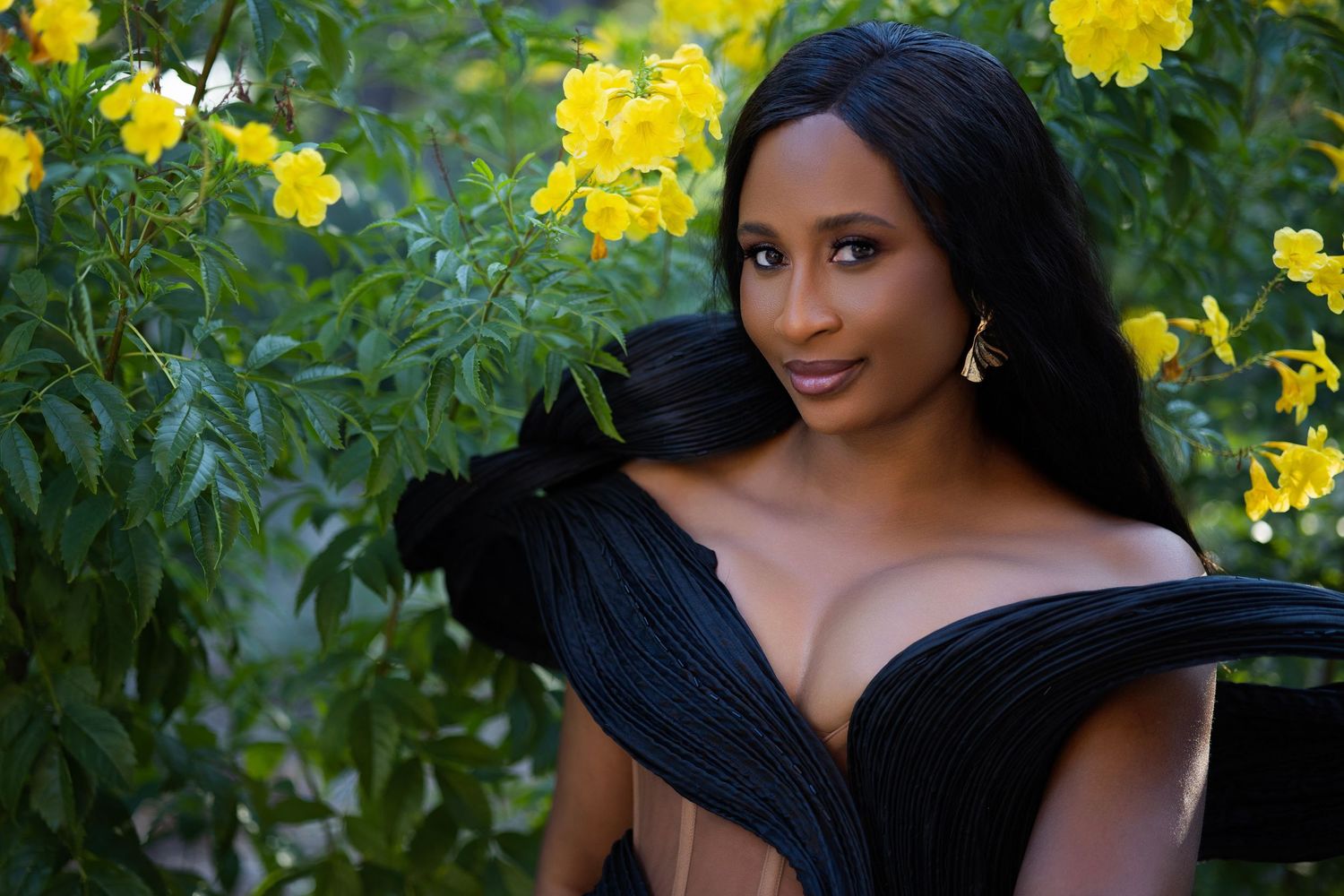 Elegant portrait with black off-shoulder dress against backdrop of yellow flowers in garden setting.