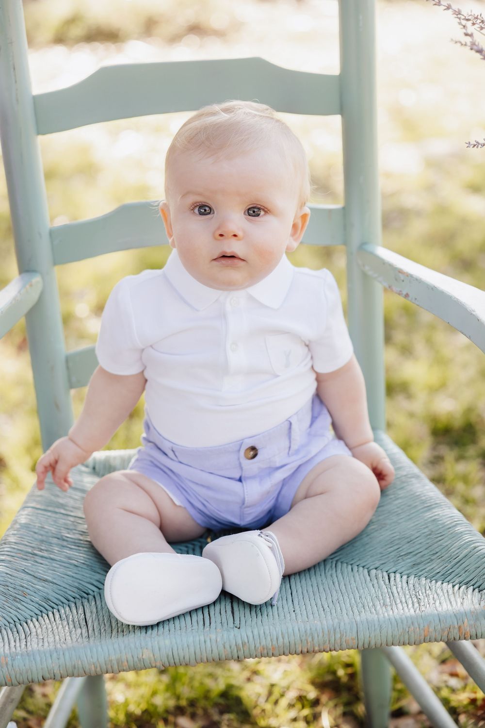 Baby wearing white shirt and blue shorts sits on turquoise chair outdoors in sunlit setting.