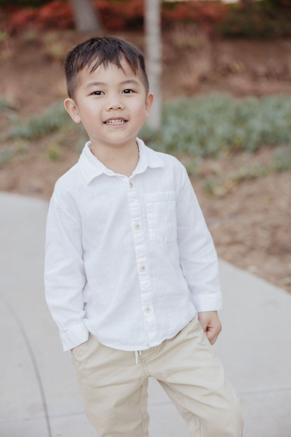 A young child wearing a white button-down shirt and khaki pants smiles while standing on a sidewalk path outdoors.