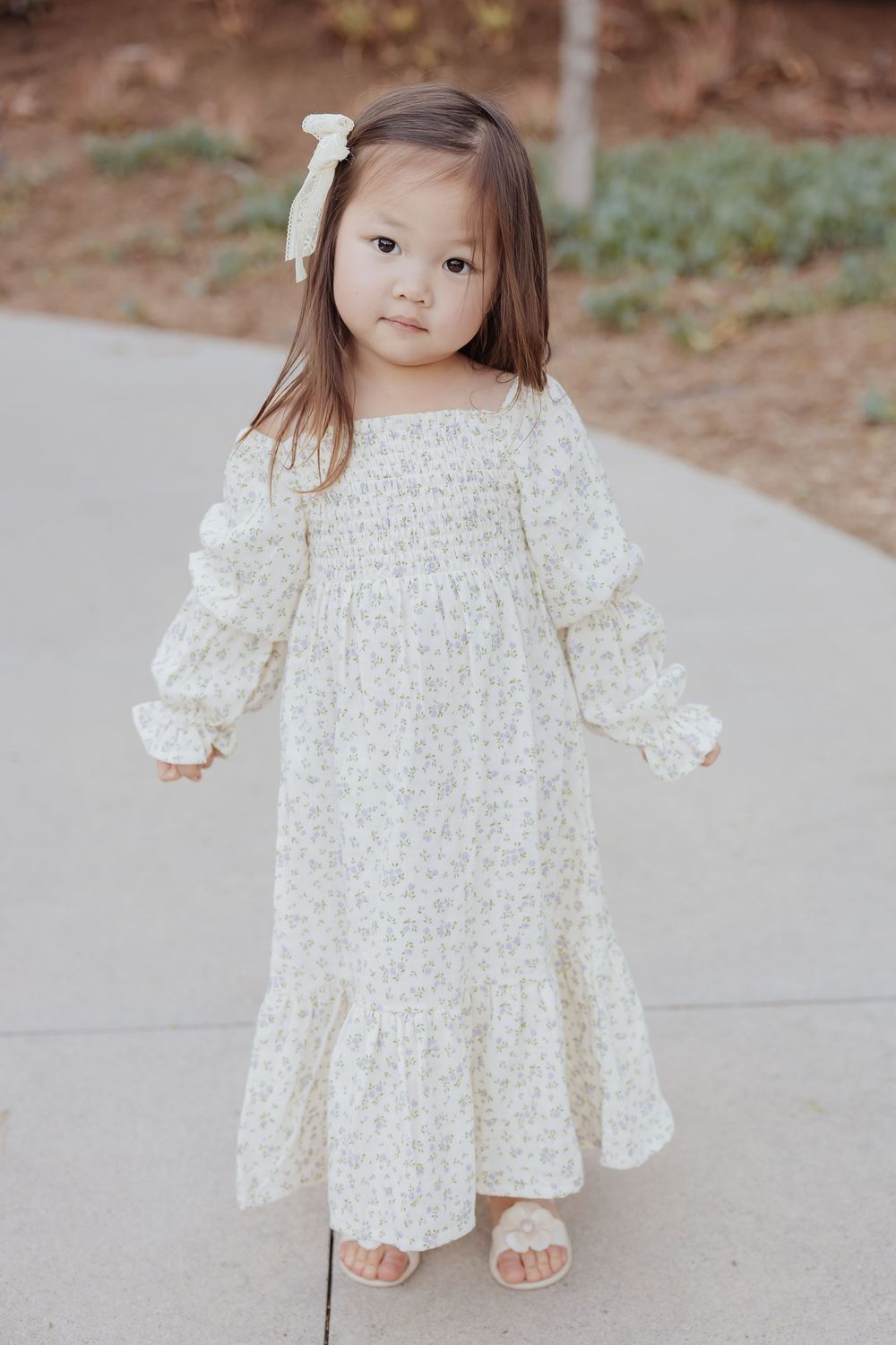 A child in a delicate cream floral dress with puff sleeves and white bow hairpiece stands on concrete path outdoors.