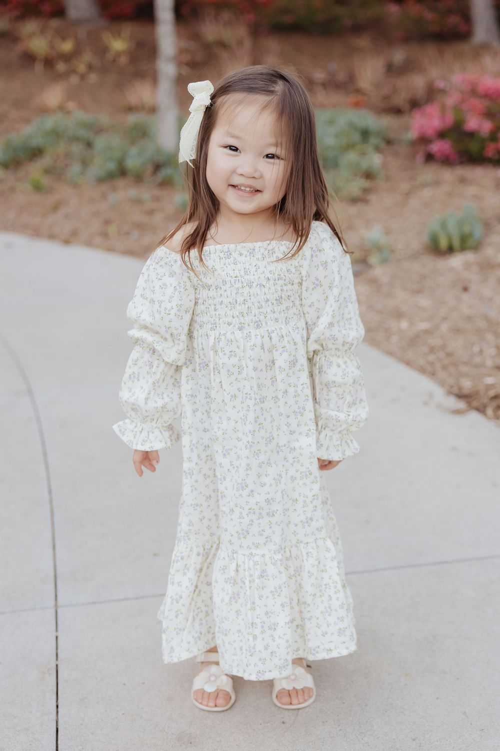 A small child wearing a white floral prairie dress with long sleeves and ruffles stands on a concrete path near garden plants.