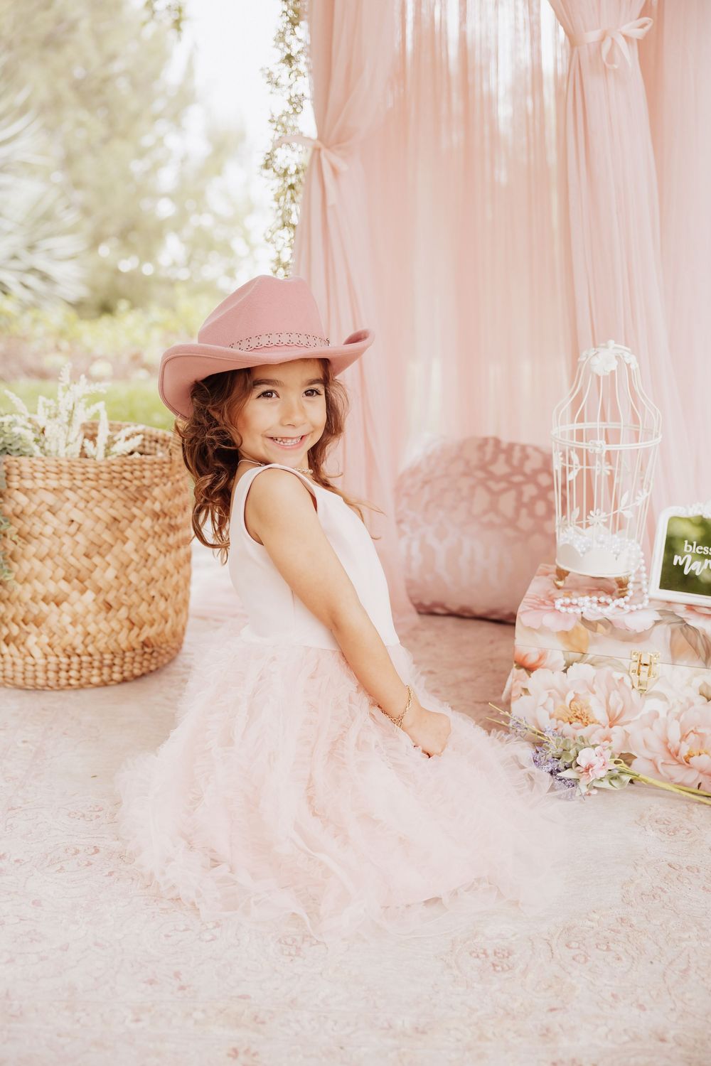 Dreamy pink photo setup with tulle curtains, decorative birdcage, straw basket and cowboy hat in soft natural lighting.