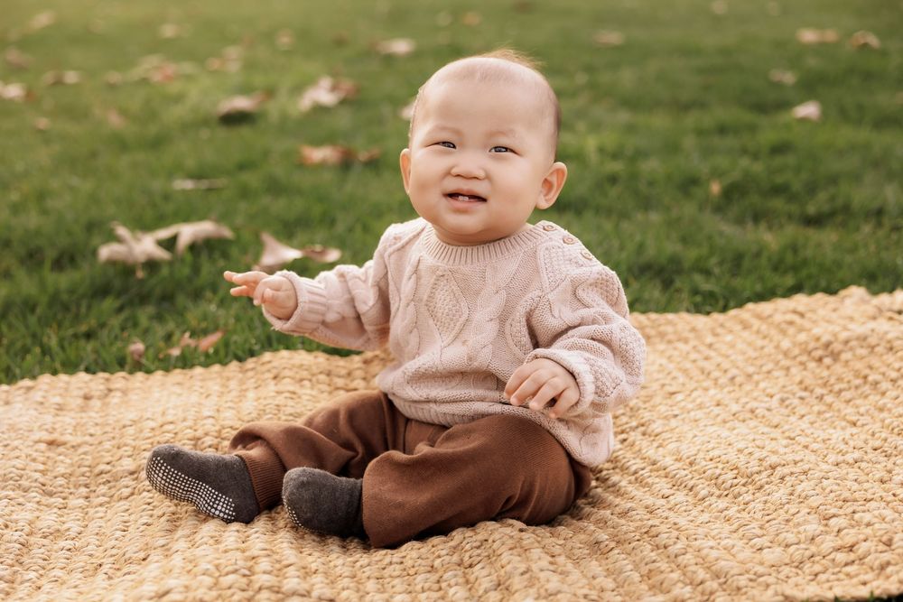 A baby wearing a light pink sweater sits and plays on a cozy tan knitted blanket outdoors.
