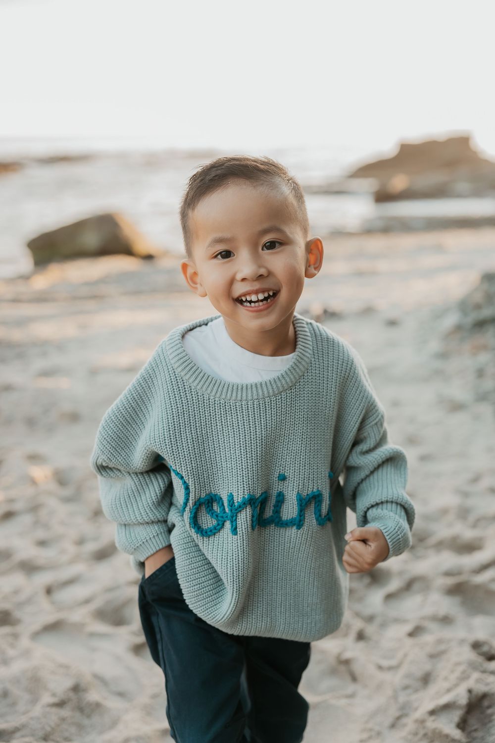 Child wearing a mint green knit sweater smiles while playing on sandy beach at sunset.