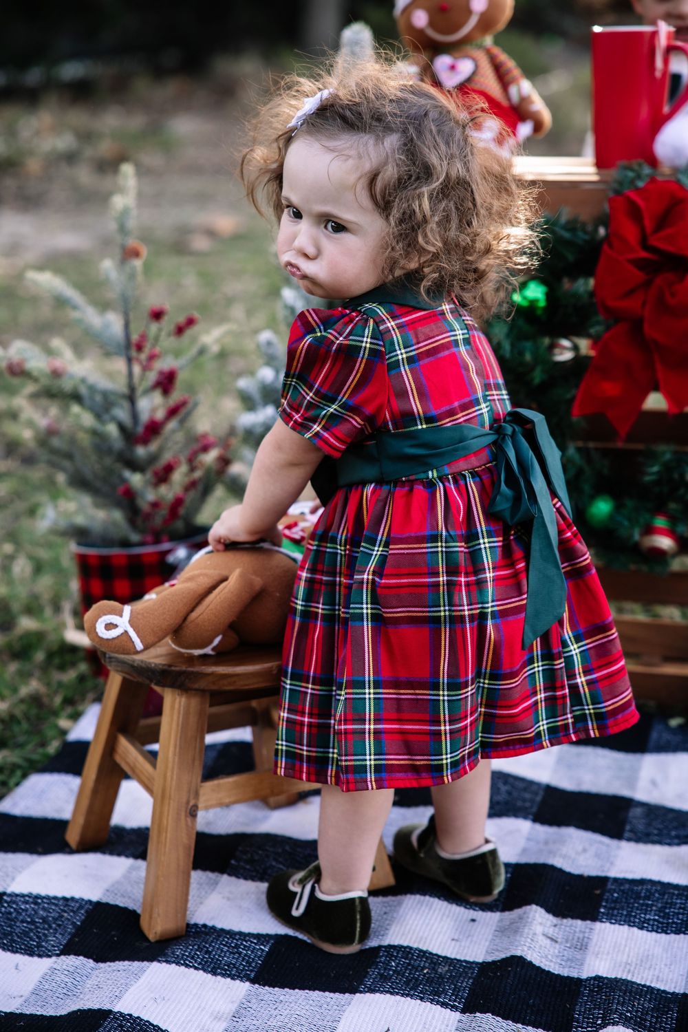 A small child in a red plaid holiday dress stands on a black and white checkered blanket near a wooden stool.