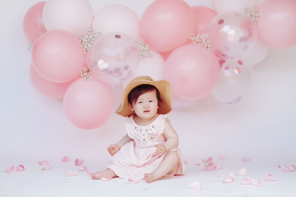Baby in sun hat sitting against pink balloon backdrop with confetti scattered on floor.