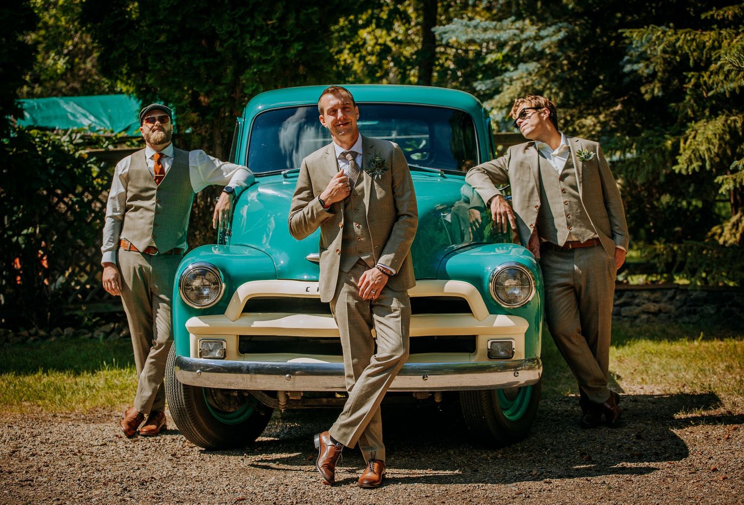 Three people in vintage suits pose in front of a classic turquoise Chevrolet pickup truck on a sunny day.