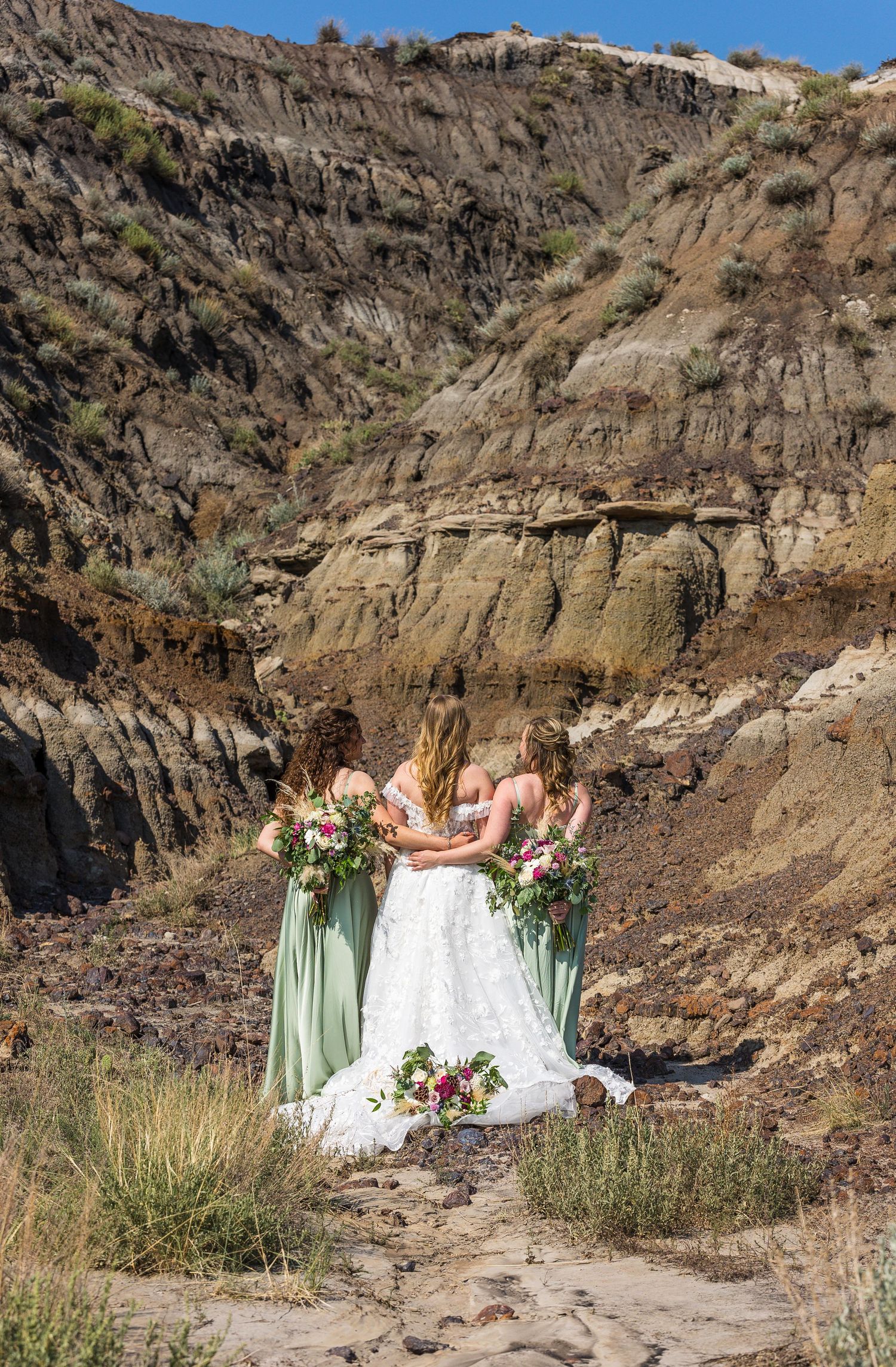 Three people in sage green and white dresses stand together against dramatic rocky cliffs on a sunny day.