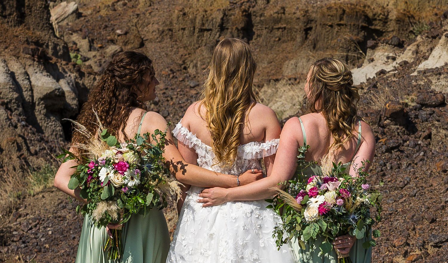 Three people in dresses stand together holding colorful floral bouquets against a rocky cliff backdrop.
