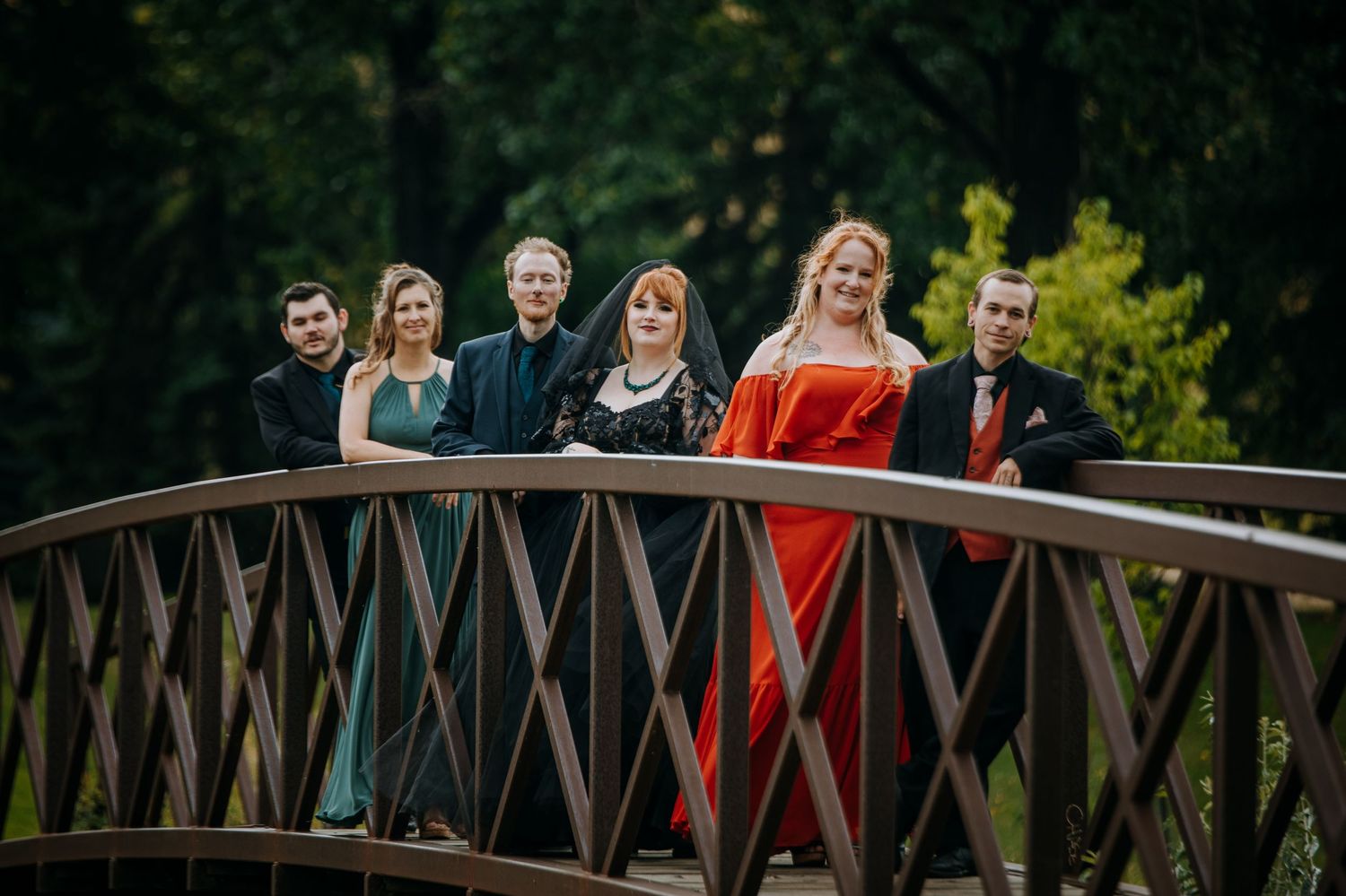 A group stands lined up on a wooden bridge in formal attire with one wearing a striking red dress against a green backdrop.