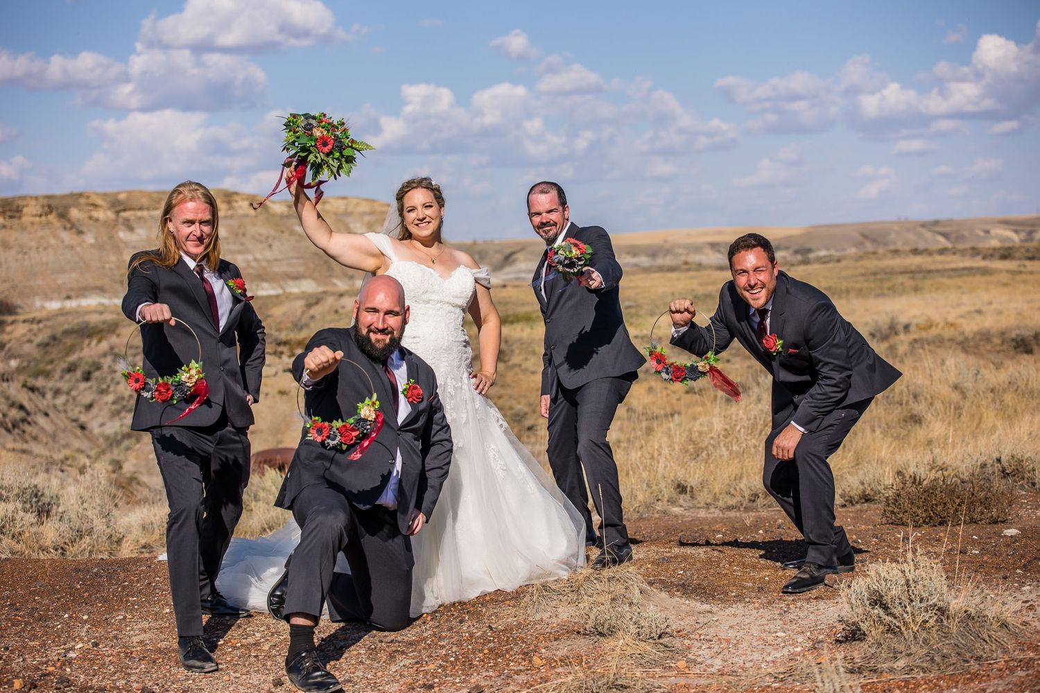 A wedding party poses playfully in an open desert landscape under blue skies with scattered clouds.