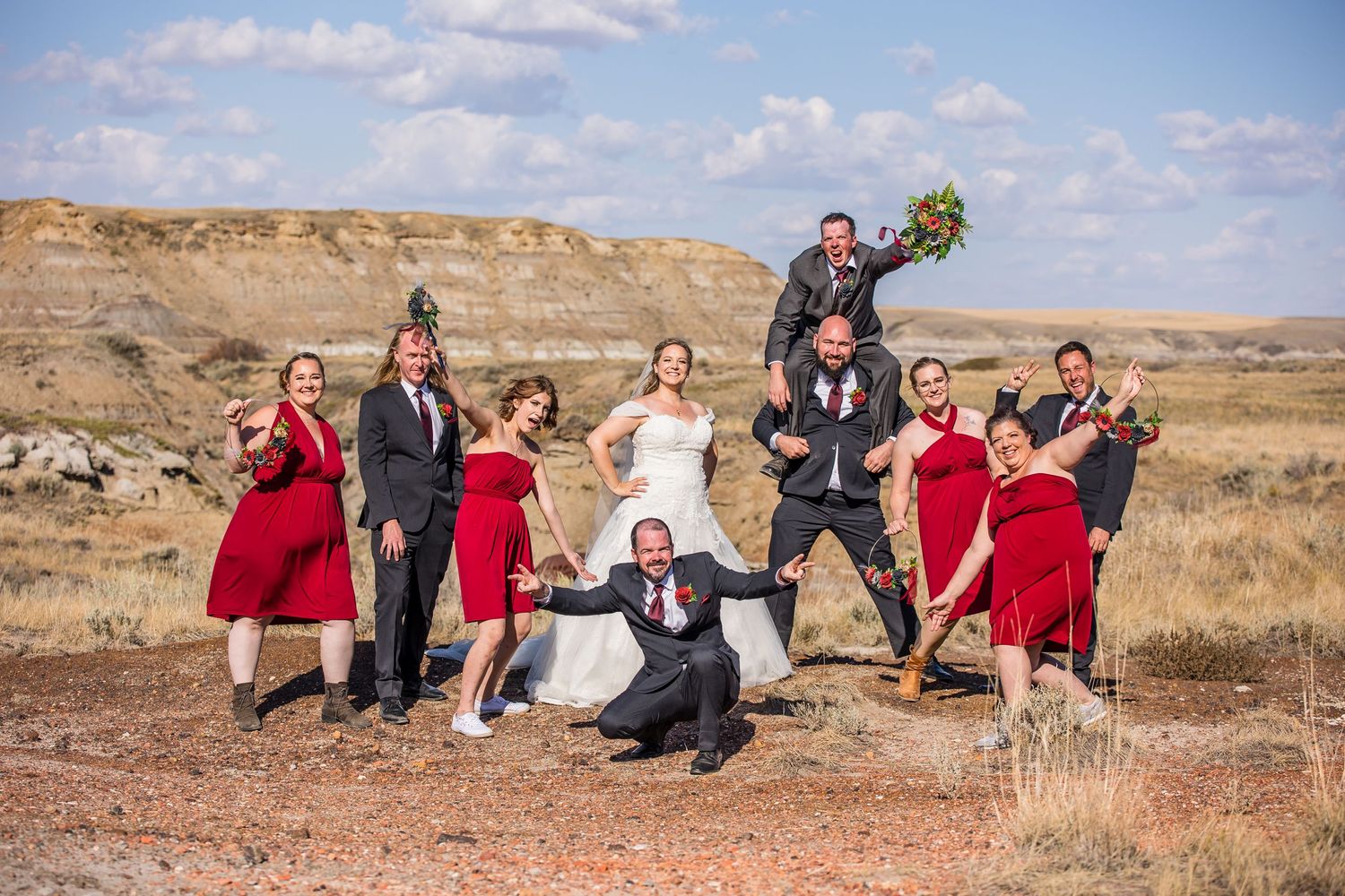 A wedding party poses playfully in bright red dresses and formal attire against a scenic desert landscape on a sunny day.