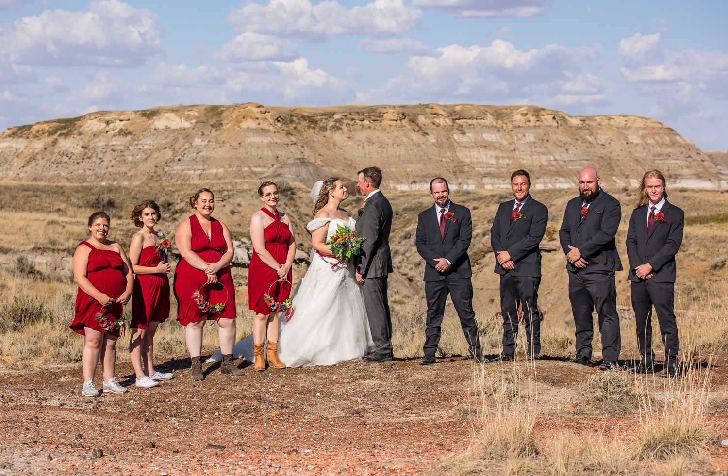 Bridal party in red dresses and dark suits pose on scenic prairie landscape with rock formations in background.