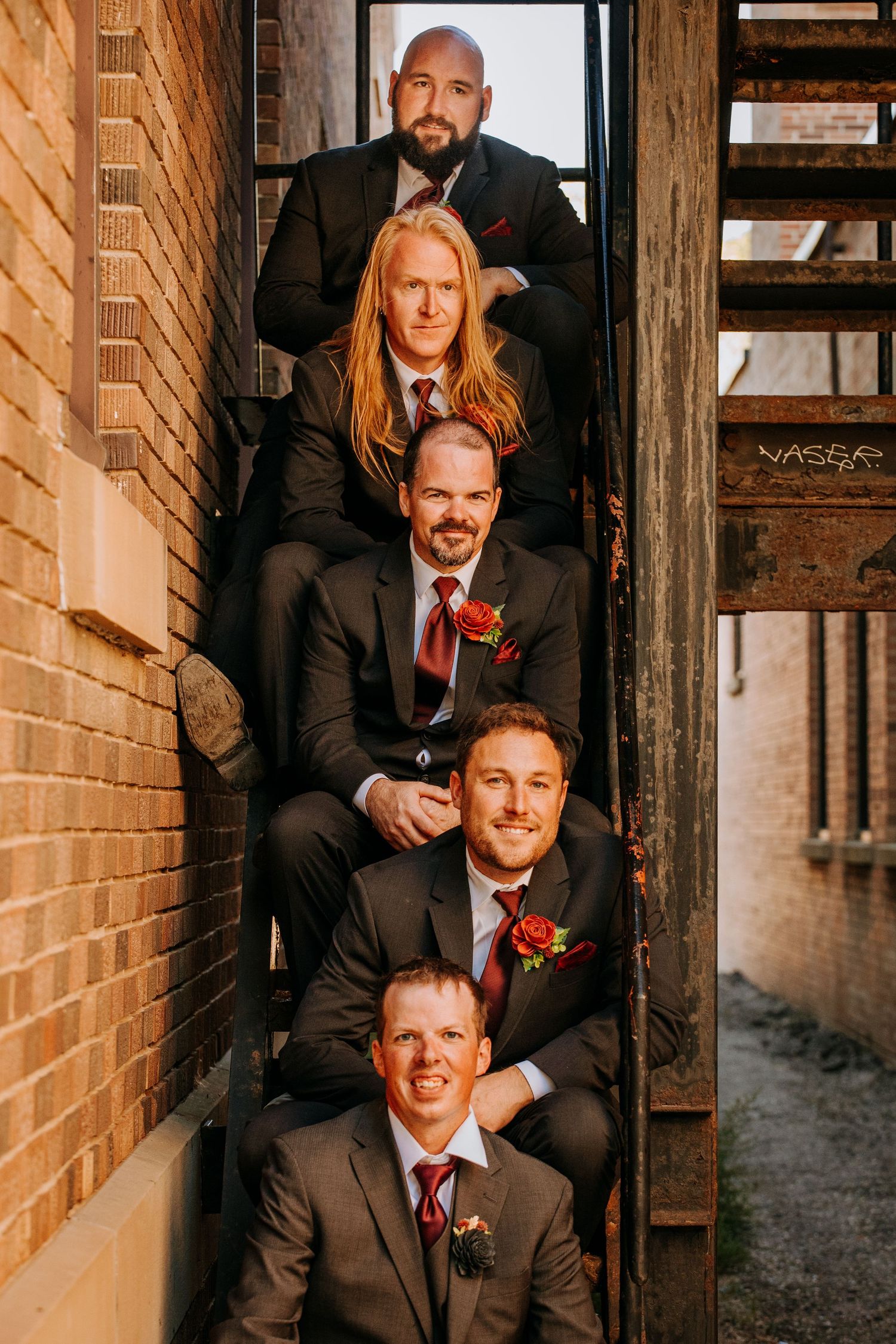Groomsmen pose together on rustic brick staircase wearing black suits with red boutonnieres during sunset lighting.