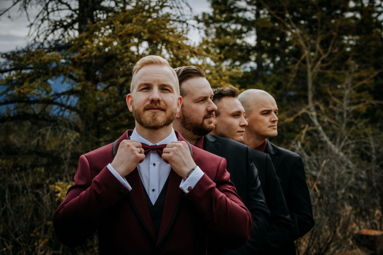 Groom in burgundy suit adjusting bow tie with groomsmen lined up behind him outdoors among trees.