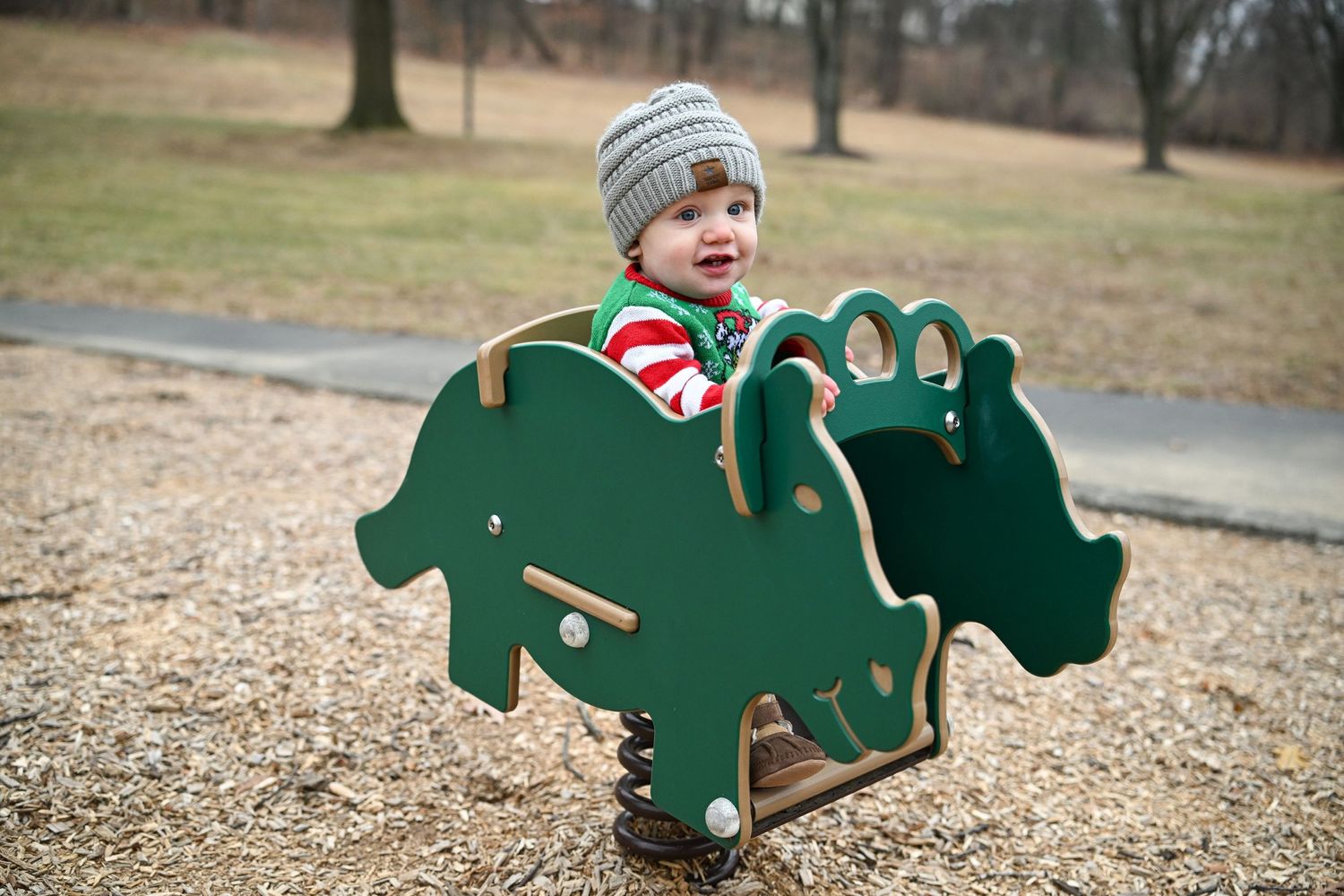 A photograph of baby boy Nolan sitting in a rocking chair shaped like a dinosaur at the park. Nolan smiles at the camera while bundled up