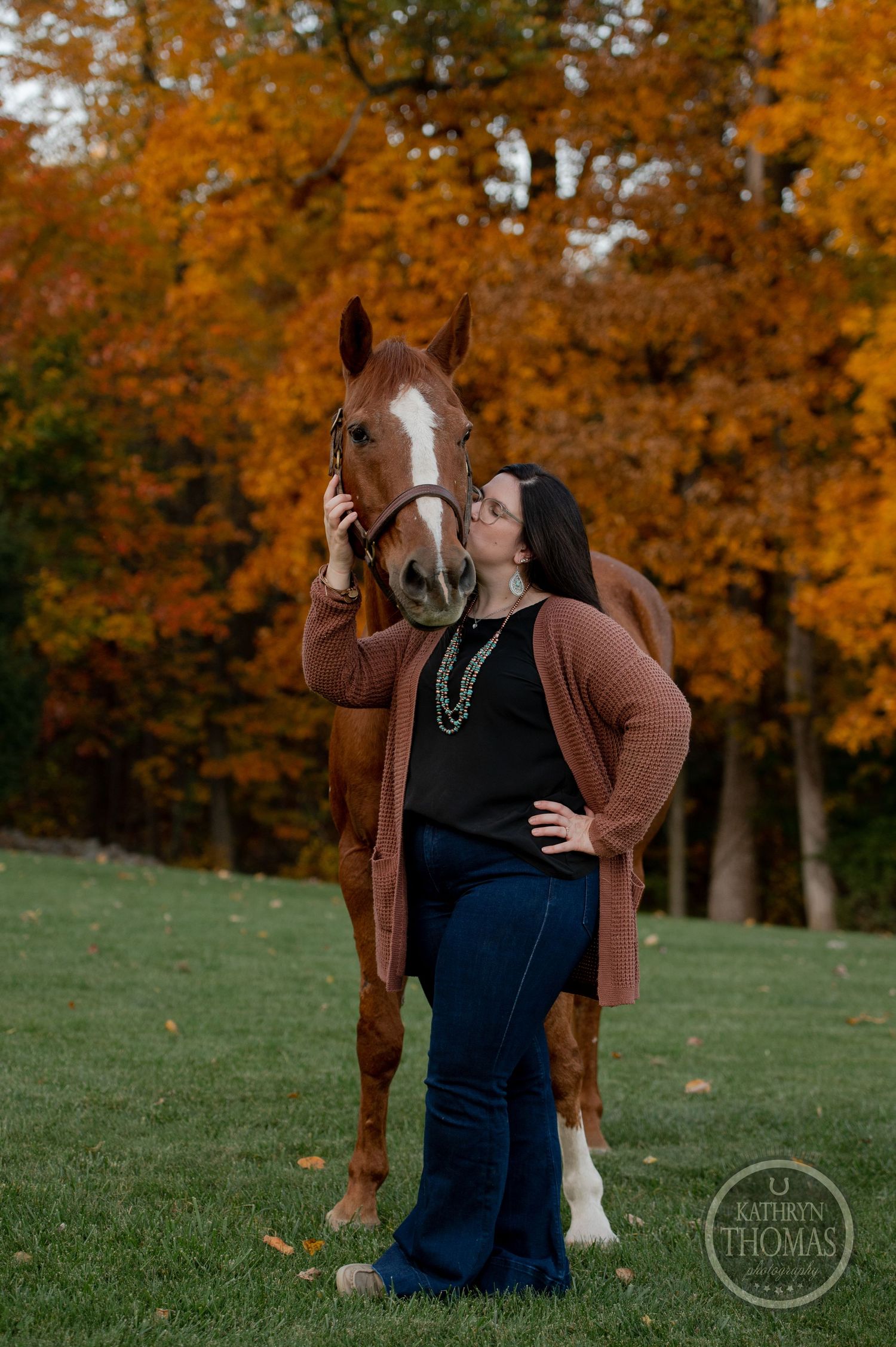 A chestnut horse with white markings poses with someone against golden autumn trees.