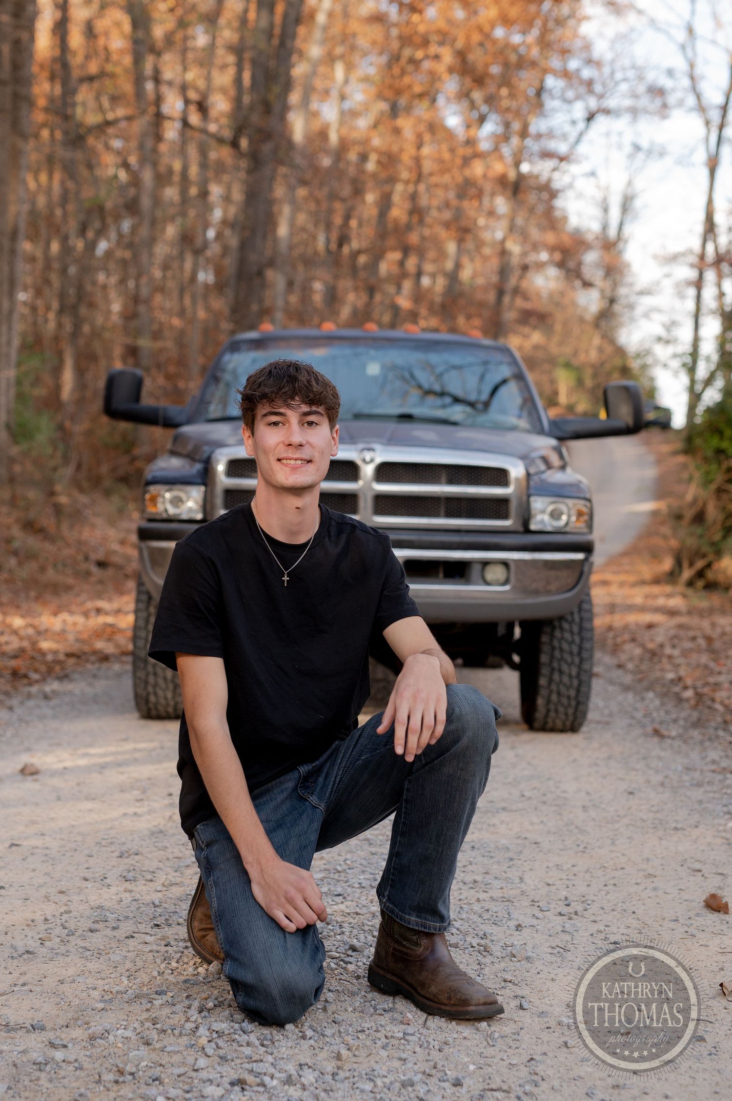 A person kneels in front of a black Dodge Ram truck on a leaf-covered forest path.