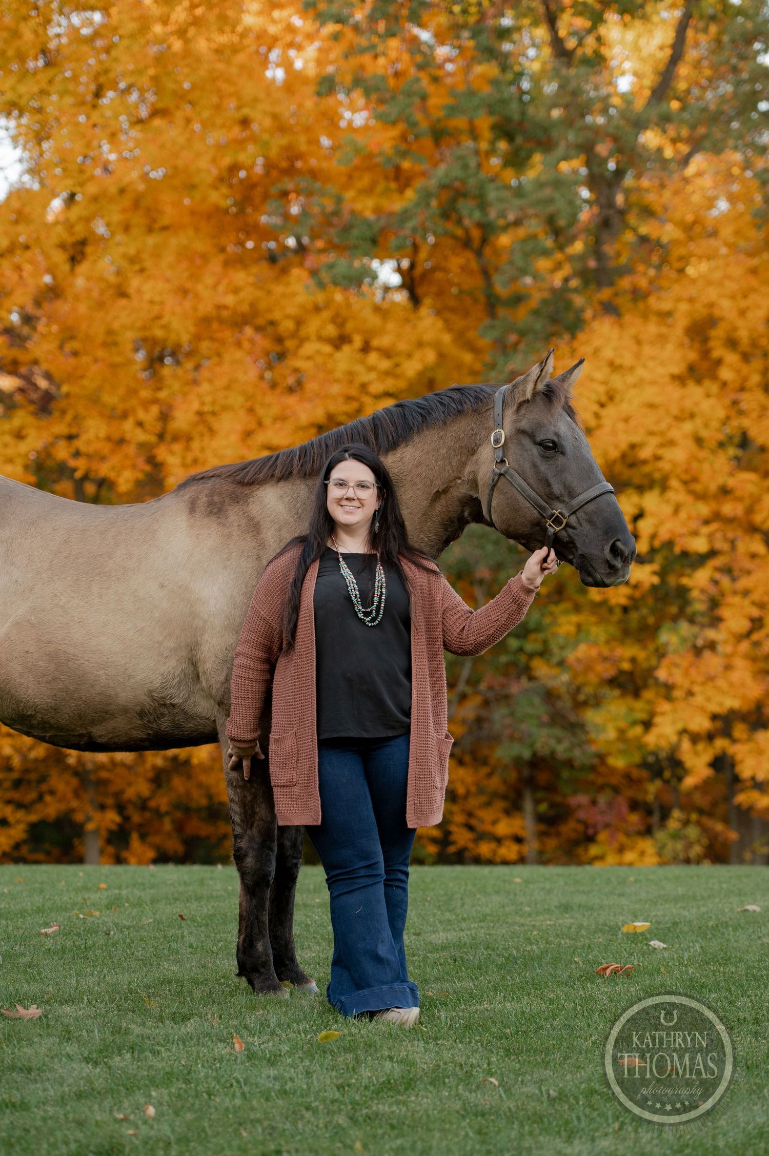 A gray horse poses with someone on a grassy hill surrounded by brilliant orange fall foliage.